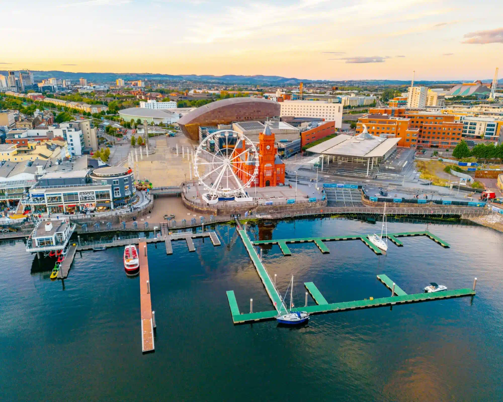 Aerial view of Cardiff Bay at sunset, with the red-brick Pierhead Building and a big wheel on the waterfront, the Wales Millennium Centre and the Senedd behind, and boats moored at the pontoons.