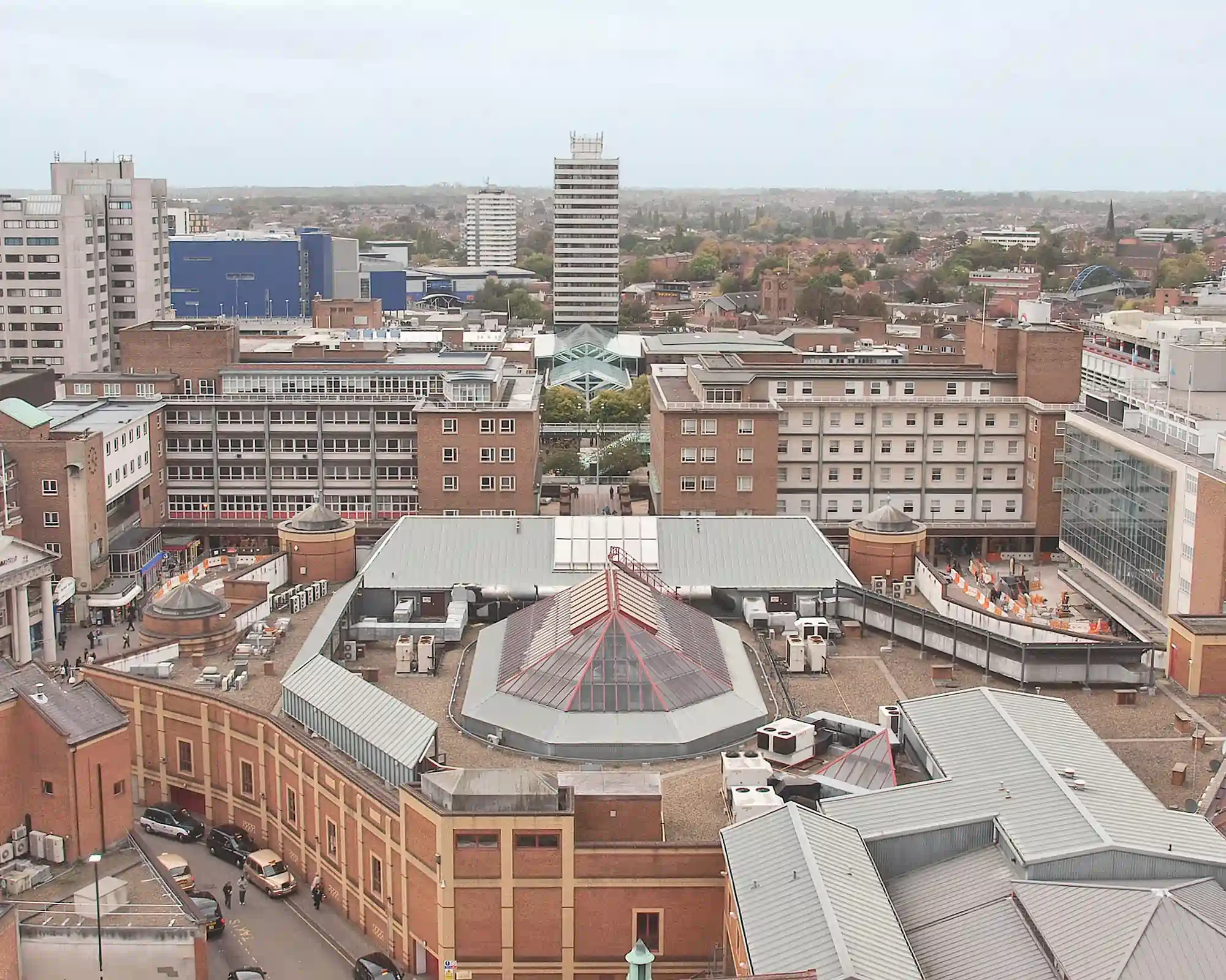 Aerial view of Coventry city centre on an overcast day, with a glass pyramid roof in the foreground, the shopping precinct and tower blocks beyond, and suburbs stretching to the horizon.