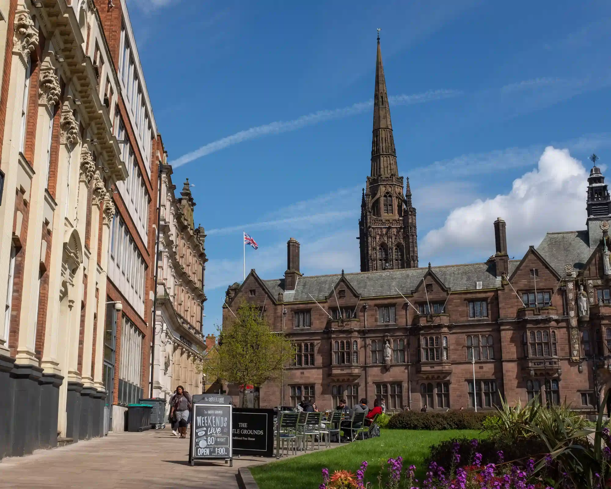 Coventry Council House, a red sandstone Tudor-style building, with the spire of the old cathedral rising behind it under a blue sky, and flower beds and outdoor seating in front.