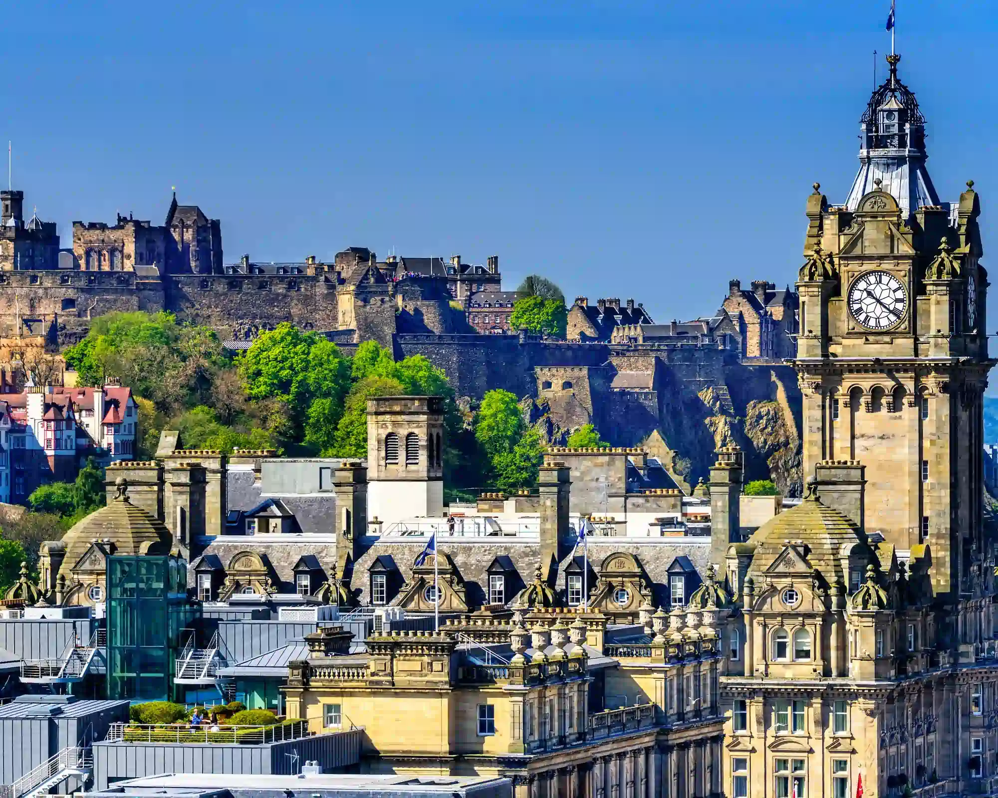 Edinburgh Castle on its rocky hill above the Old Town, with the Balmoral Hotel's clock tower in the foreground under a clear blue sky.