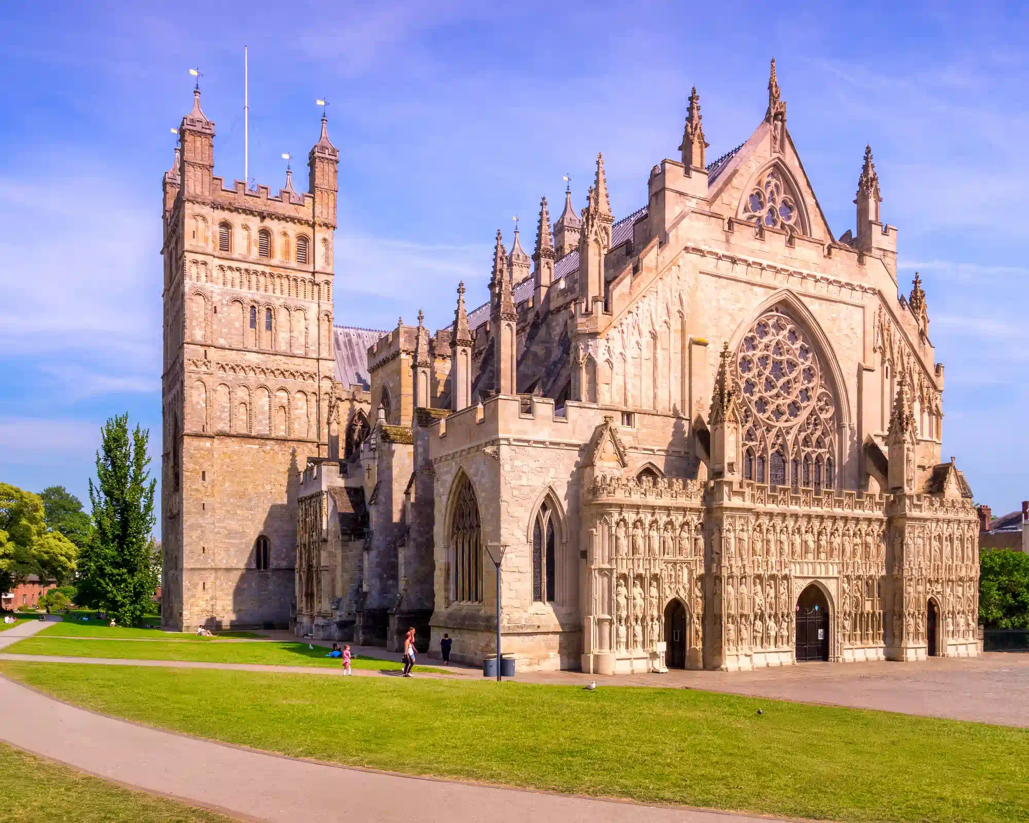 The west front of Exeter Cathedral on a sunny day, with its carved stone screen of statues, large rose window and Norman tower, set behind green lawns.