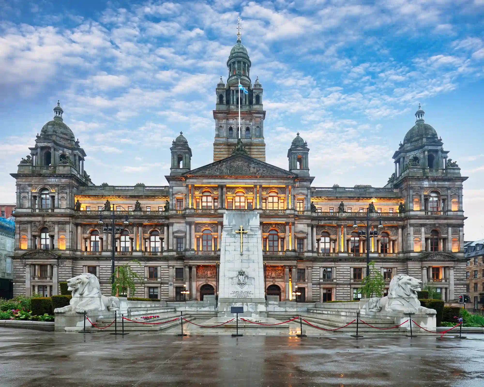 Glasgow City Chambers in George Square, lit up at dusk, with the cenotaph and two stone lions in front under a cloudy blue sky.