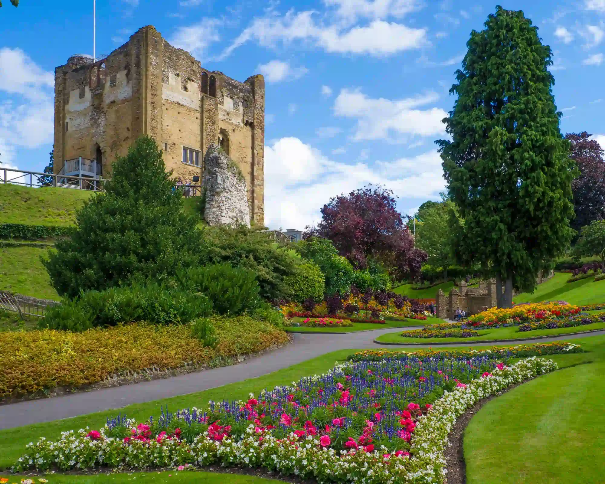 The Norman keep of Guildford Castle on a grassy mound above colourful flower beds, winding paths and tall trees in the castle gardens, under a blue sky.