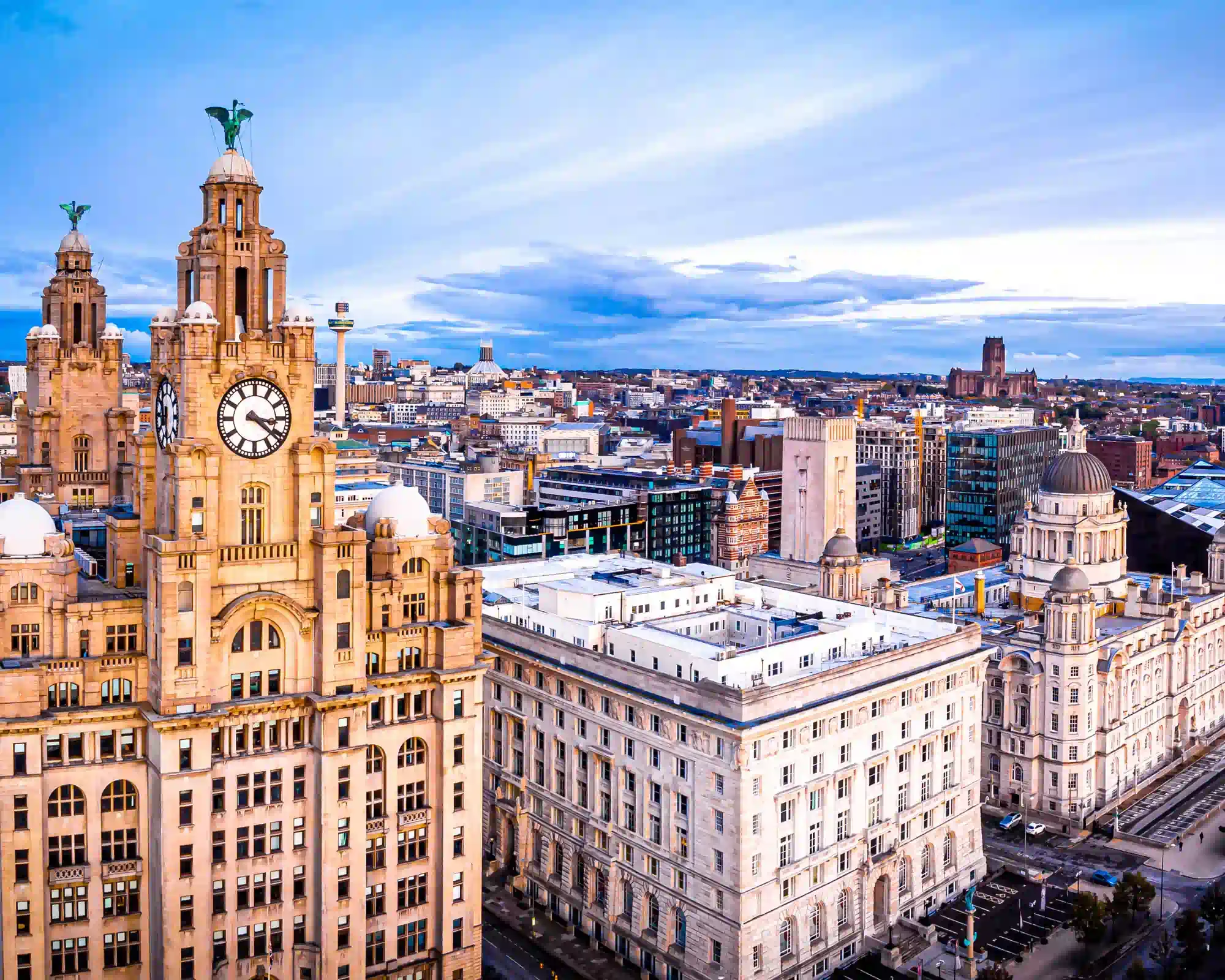 Aerial view of Liverpool's waterfront, with the Royal Liver Building's clock towers and Liver Birds beside the Cunard and Port of Liverpool buildings, and the city stretching to the cathedral beyond.