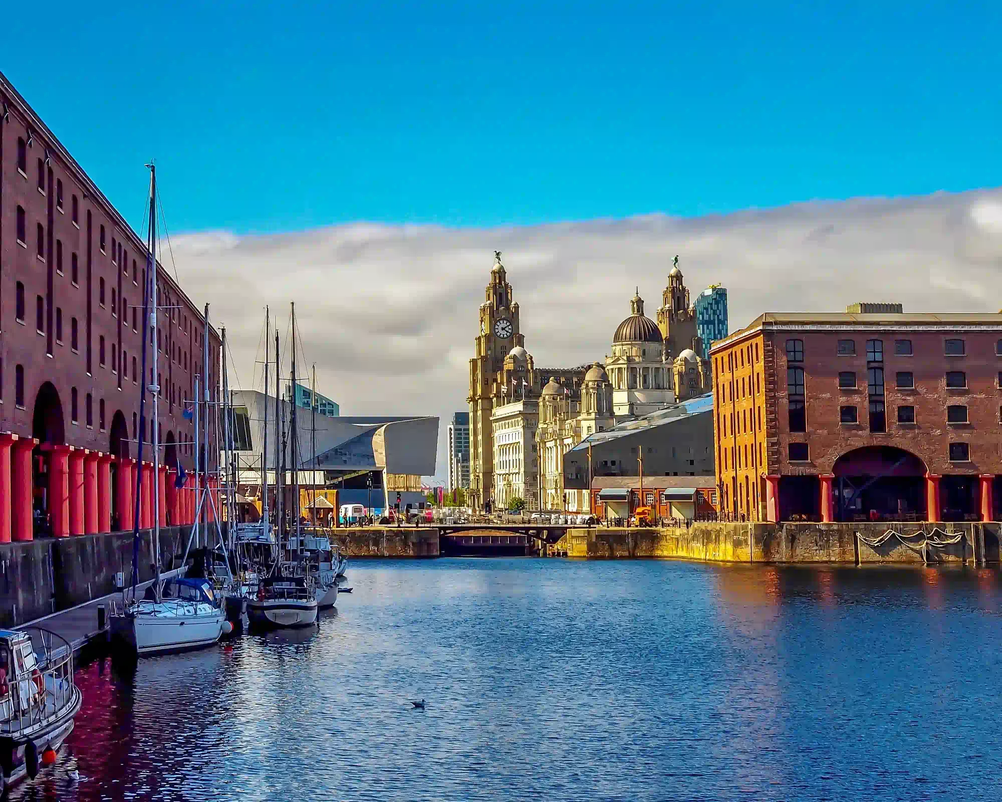 Royal Albert Dock in Liverpool on a sunny day, with red-columned brick warehouses and moored boats along the water, and the Royal Liver Building beyond.