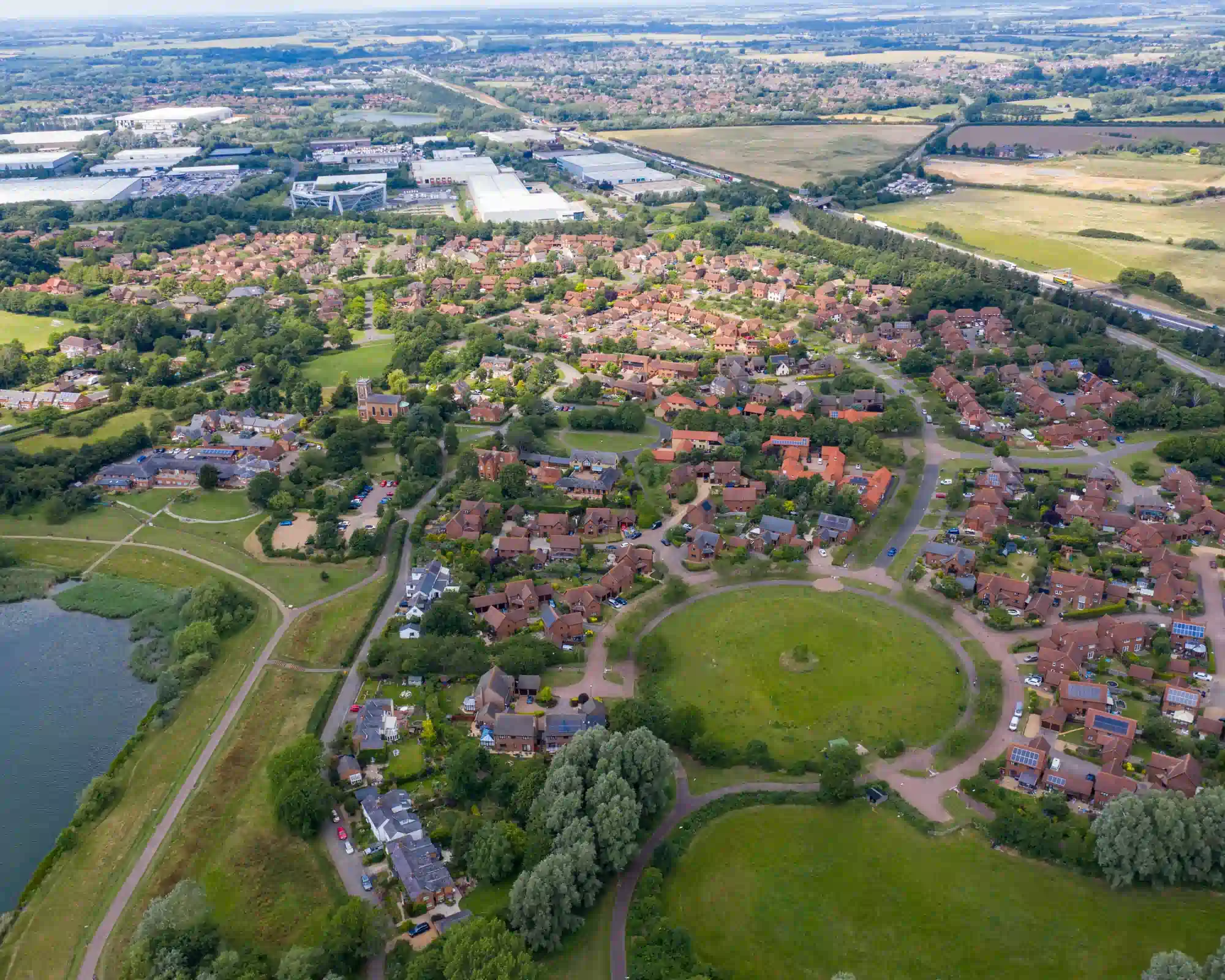 Aerial view of a Milton Keynes neighbourhood on a sunny day, with red-brick houses around a circular green, a lake and paths on one side, and business parks and fields beyond.