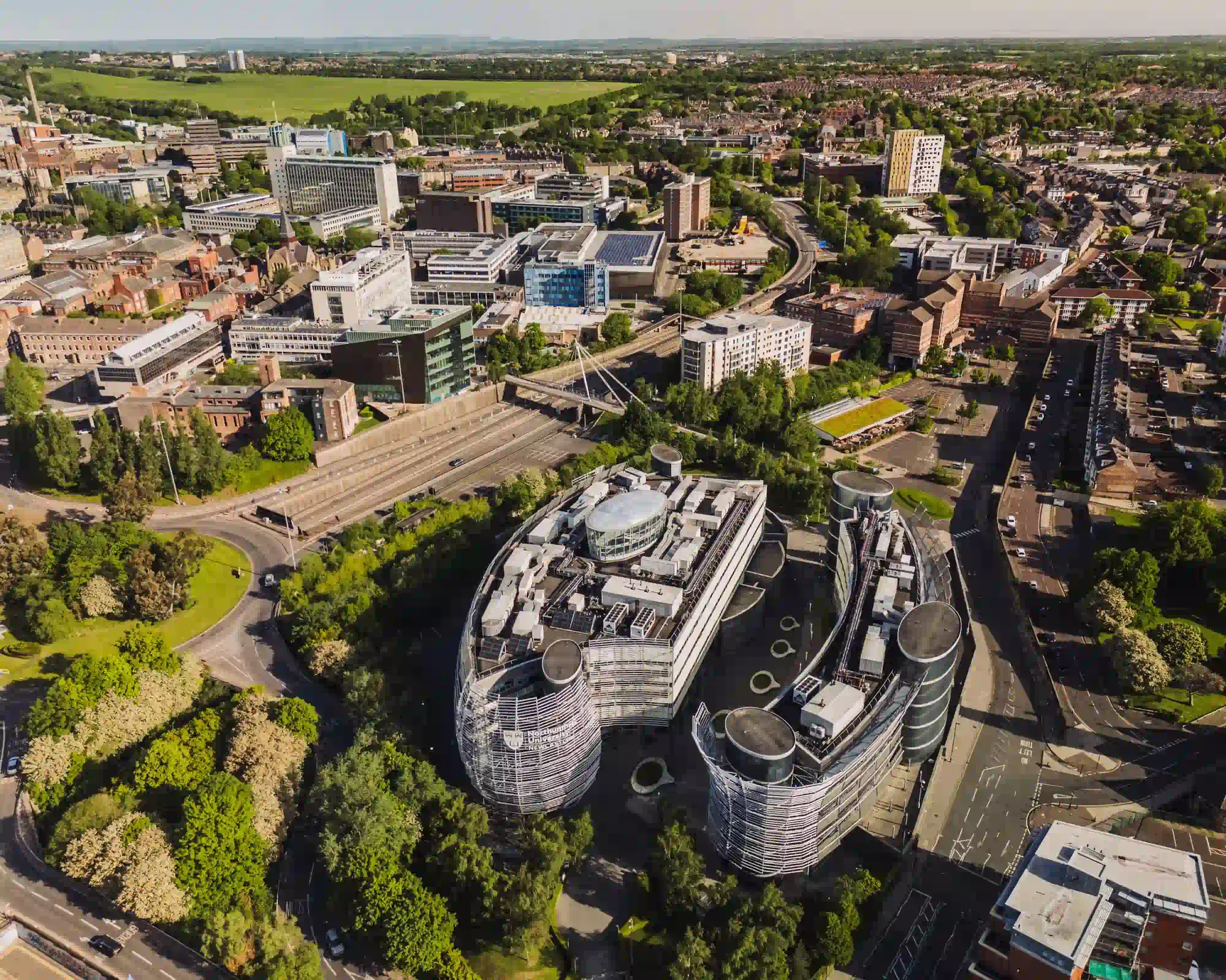 Aerial view of Newcastle on a sunny day, with Northumbria University's curved glass and steel buildings among trees, and the city centre and Town Moor beyond.