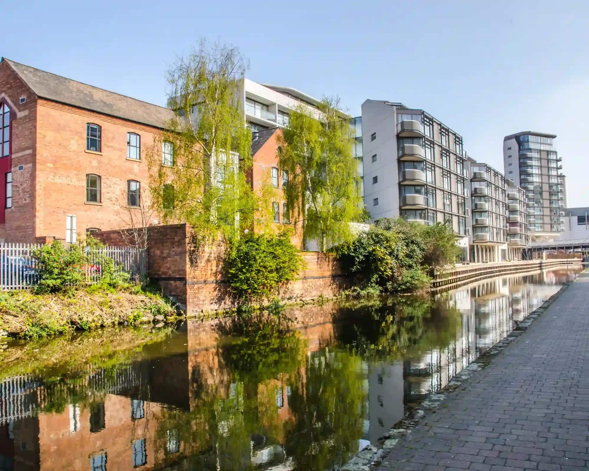 The Nottingham Canal on a sunny day, with a red-brick warehouse and modern apartment blocks reflected in the water beside a paved towpath.