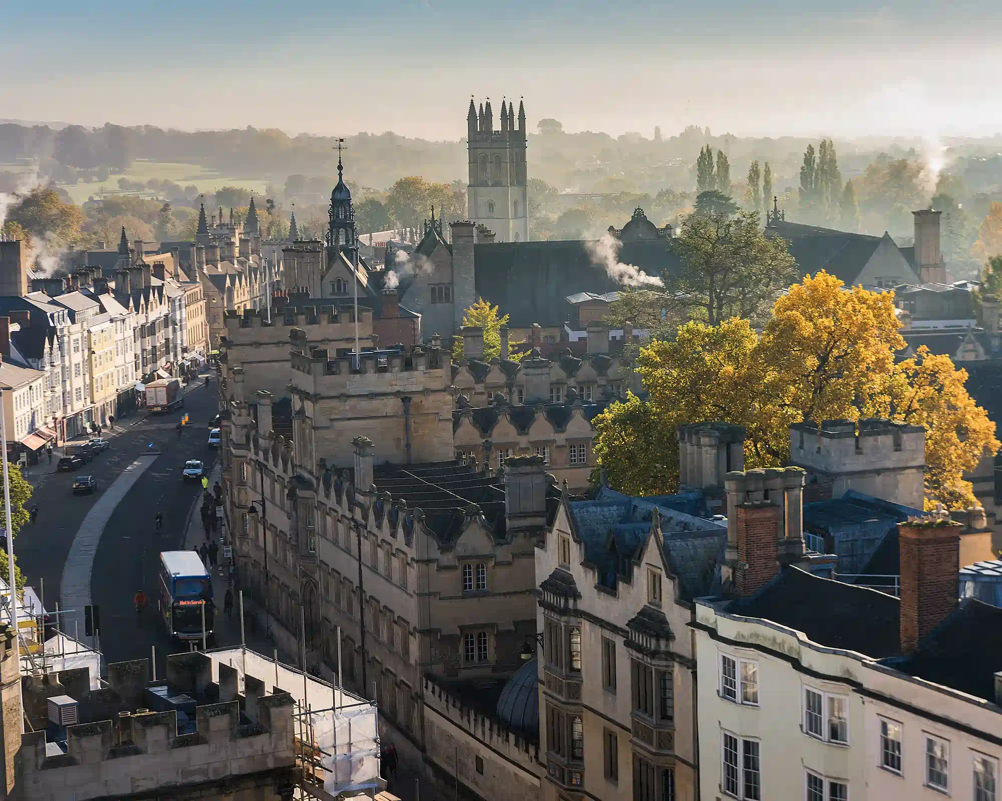 Oxford's High Street on a misty autumn morning, with Magdalen College's tower rising above stone college buildings, a golden tree and parkland beyond.