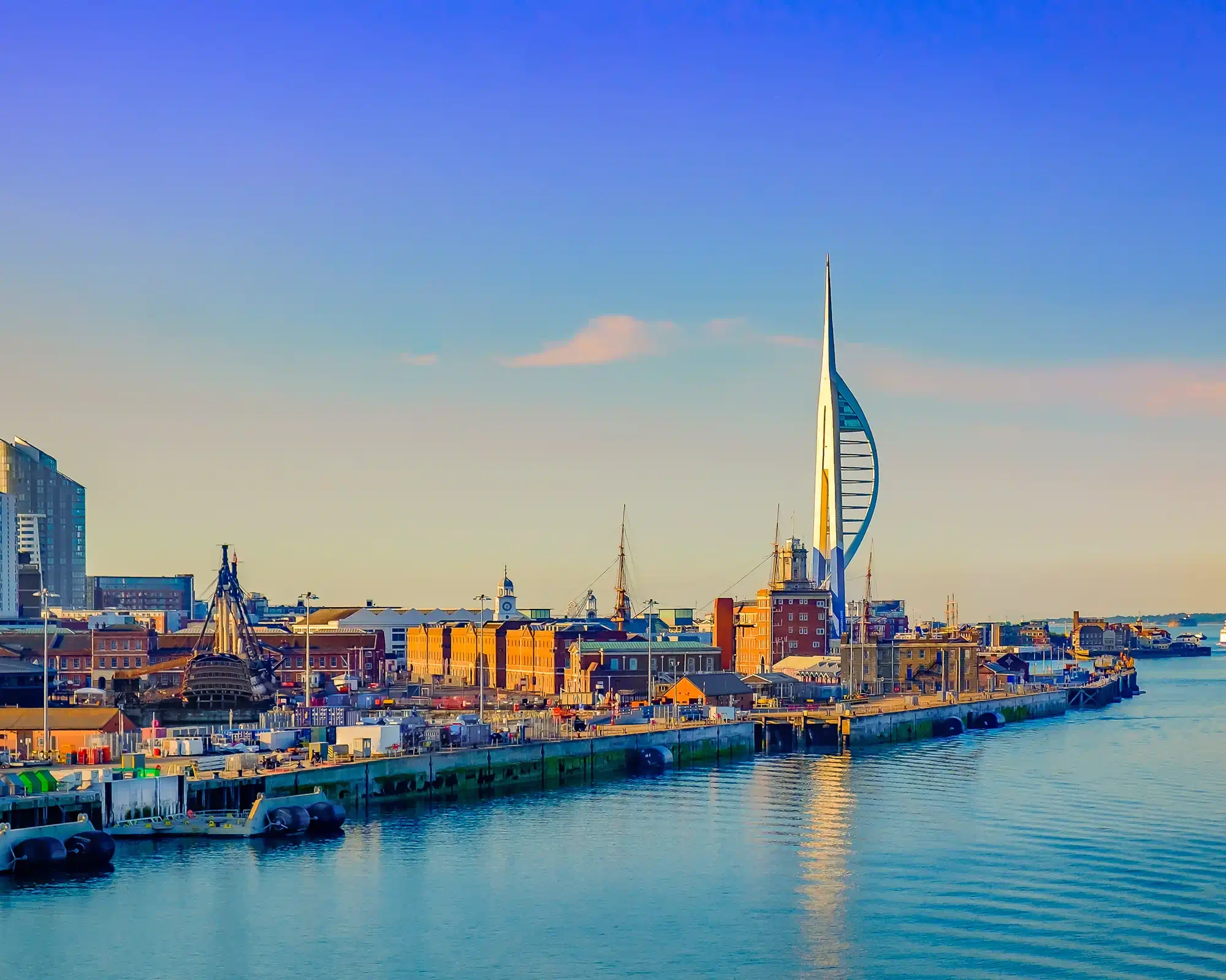 Portsmouth's Spinnaker Tower glowing in golden evening light above the Historic Dockyard, with HMS Victory's masts and red-brick buildings reflected in the harbour.