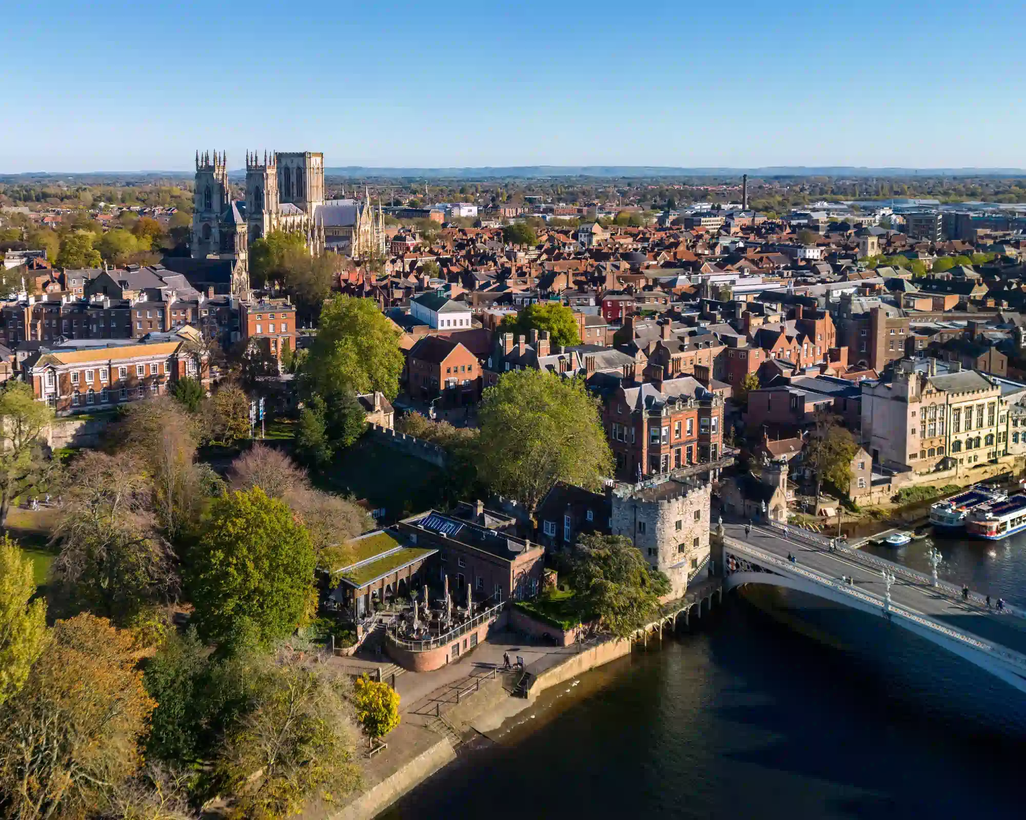 Aerial view of York on a sunny autumn day, with York Minster rising above red-brick rooftops, and Lendal Bridge crossing the River Ouse past moored tour boats.