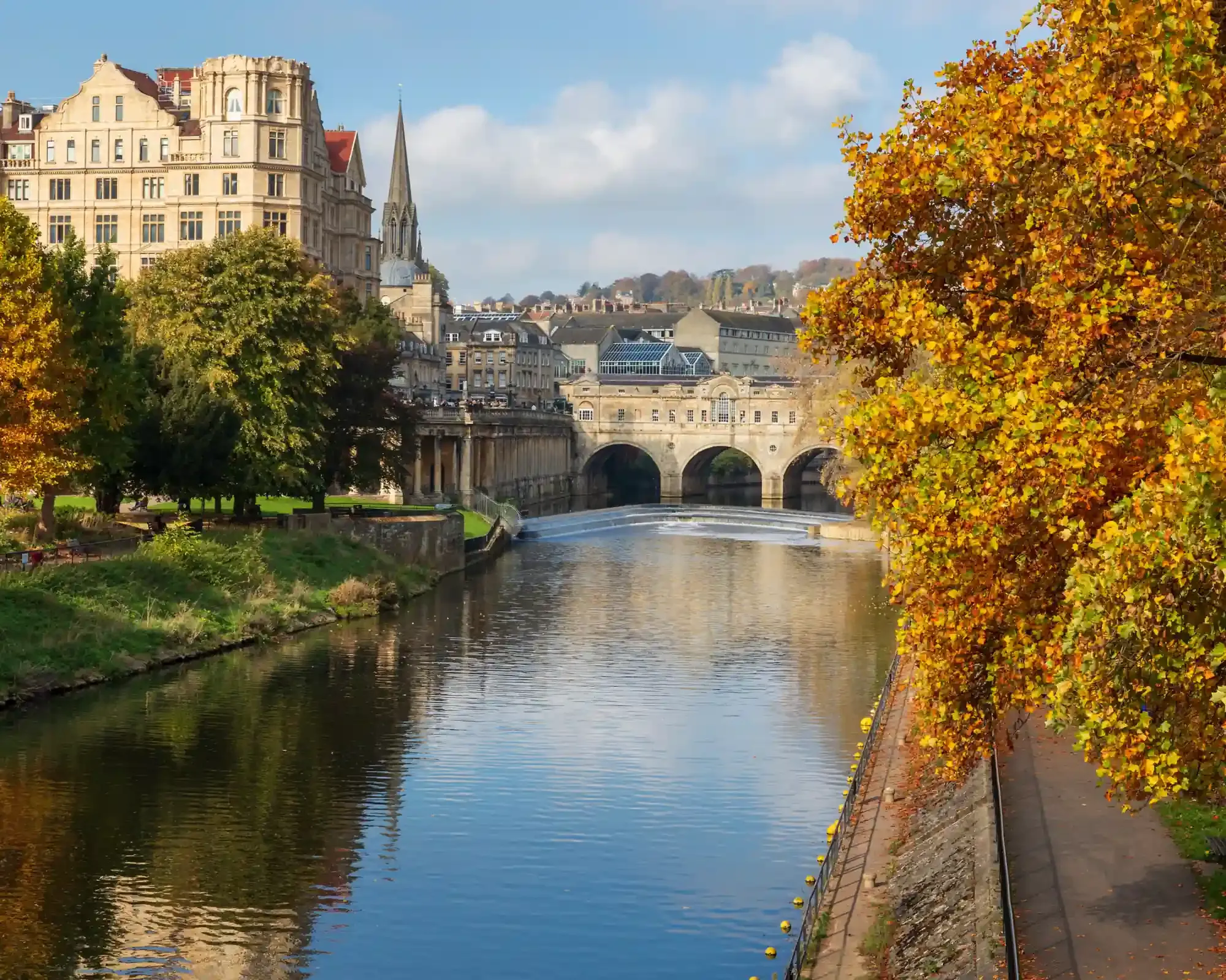 Pulteney Bridge and its weir on the River Avon in Bath, framed by autumn trees and honey-coloured stone buildings.