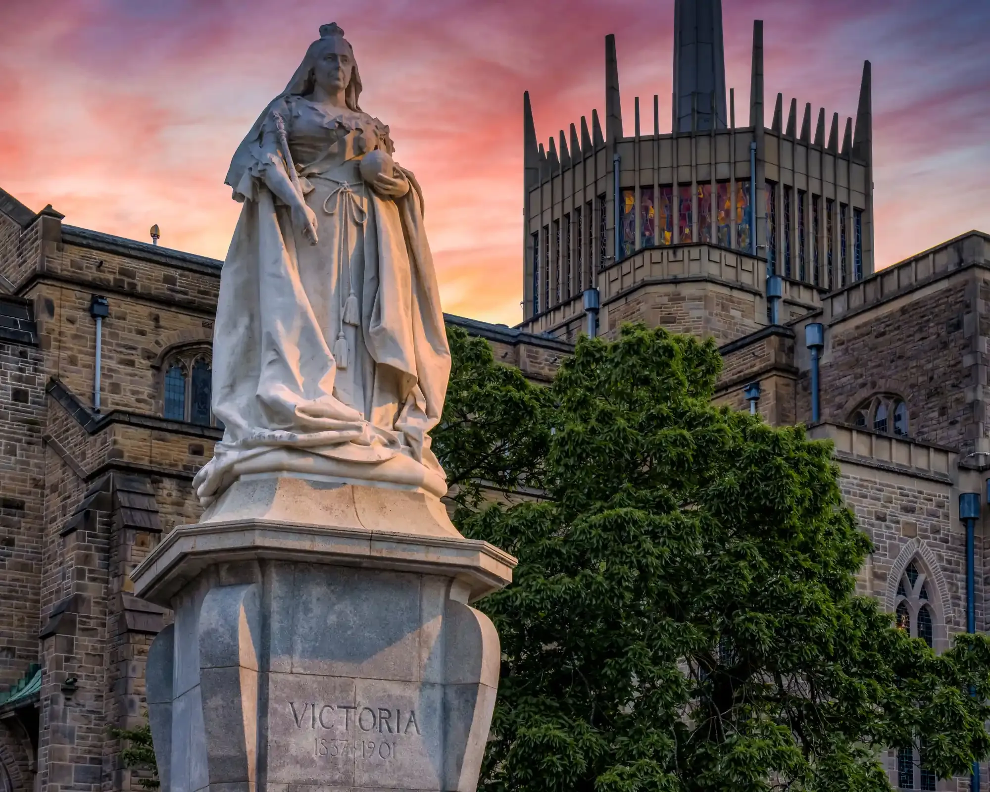 Queen Victoria statue outside Blackburn Cathedral, with the cathedral's lantern tower against a pink sunset sky.