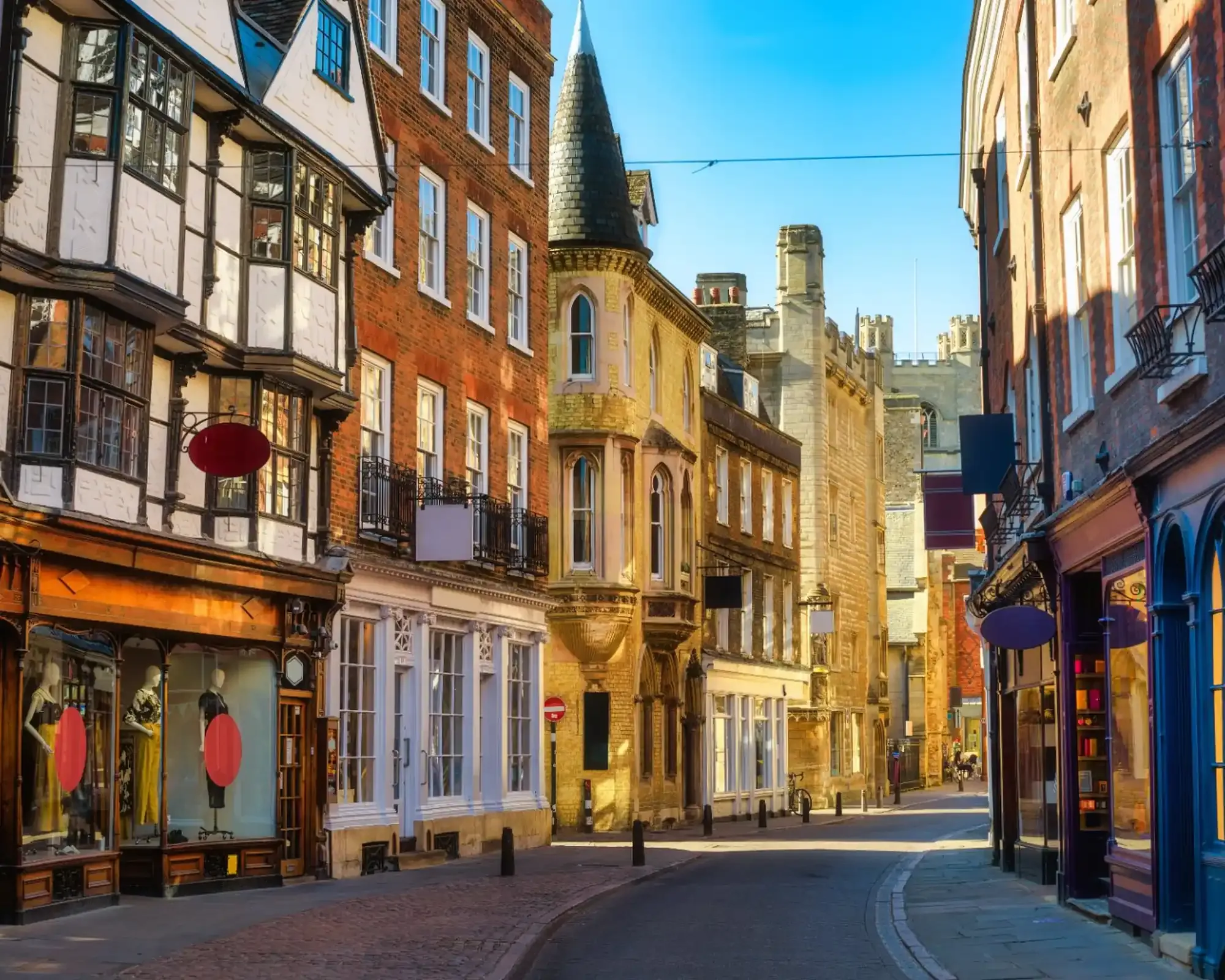Historic shopfronts and a turreted building on a sunlit street in central Cambridge, with college towers at the far end.