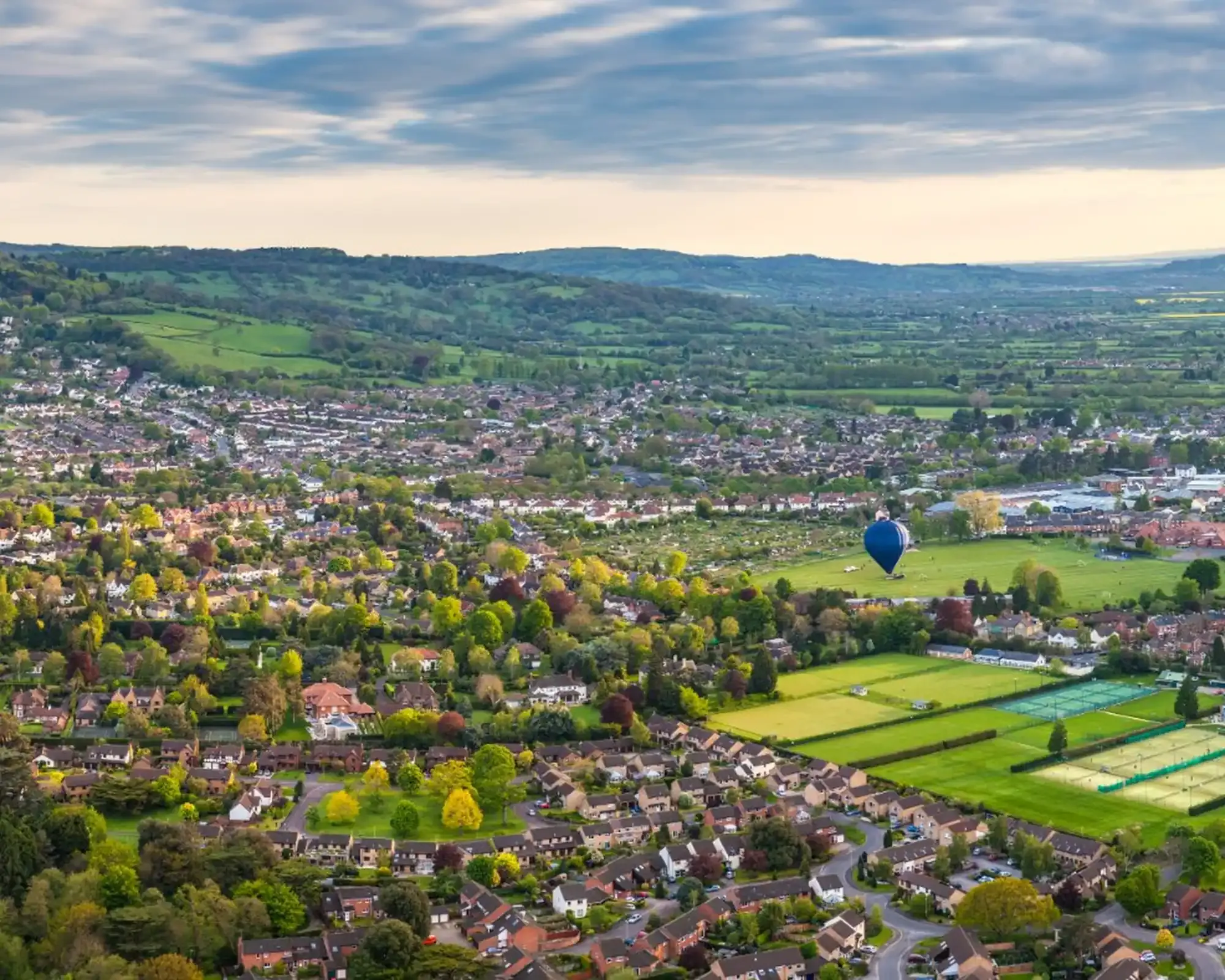 Aerial view over Cheltenham's leafy suburbs and playing fields, with a blue hot air balloon landing on the grass and Cotswold hills in the distance.