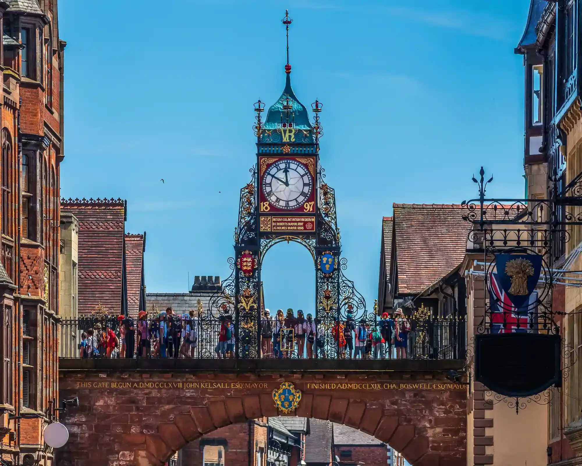 The Victorian Eastgate Clock above the sandstone arch of Chester's city walls, with people looking out from the walkway on the walls.