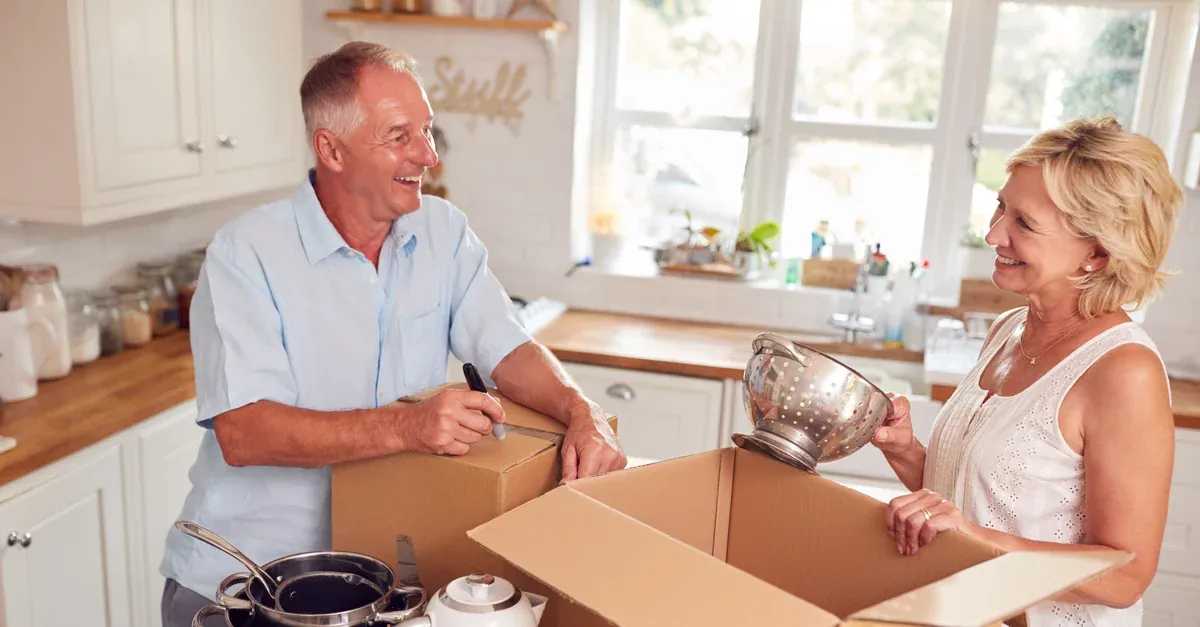 An couple in their 60s in a bright kitchen packing crockery into boxes and taping them up.