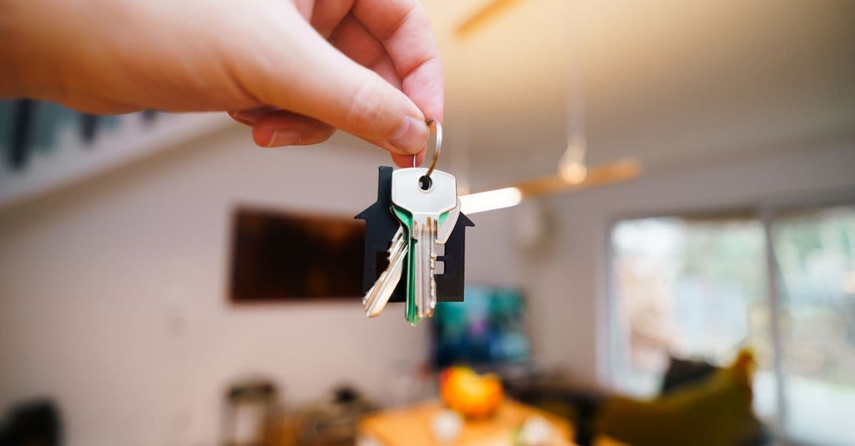 A hand holds keys with a house keychain inside a modern, well-lit living space.