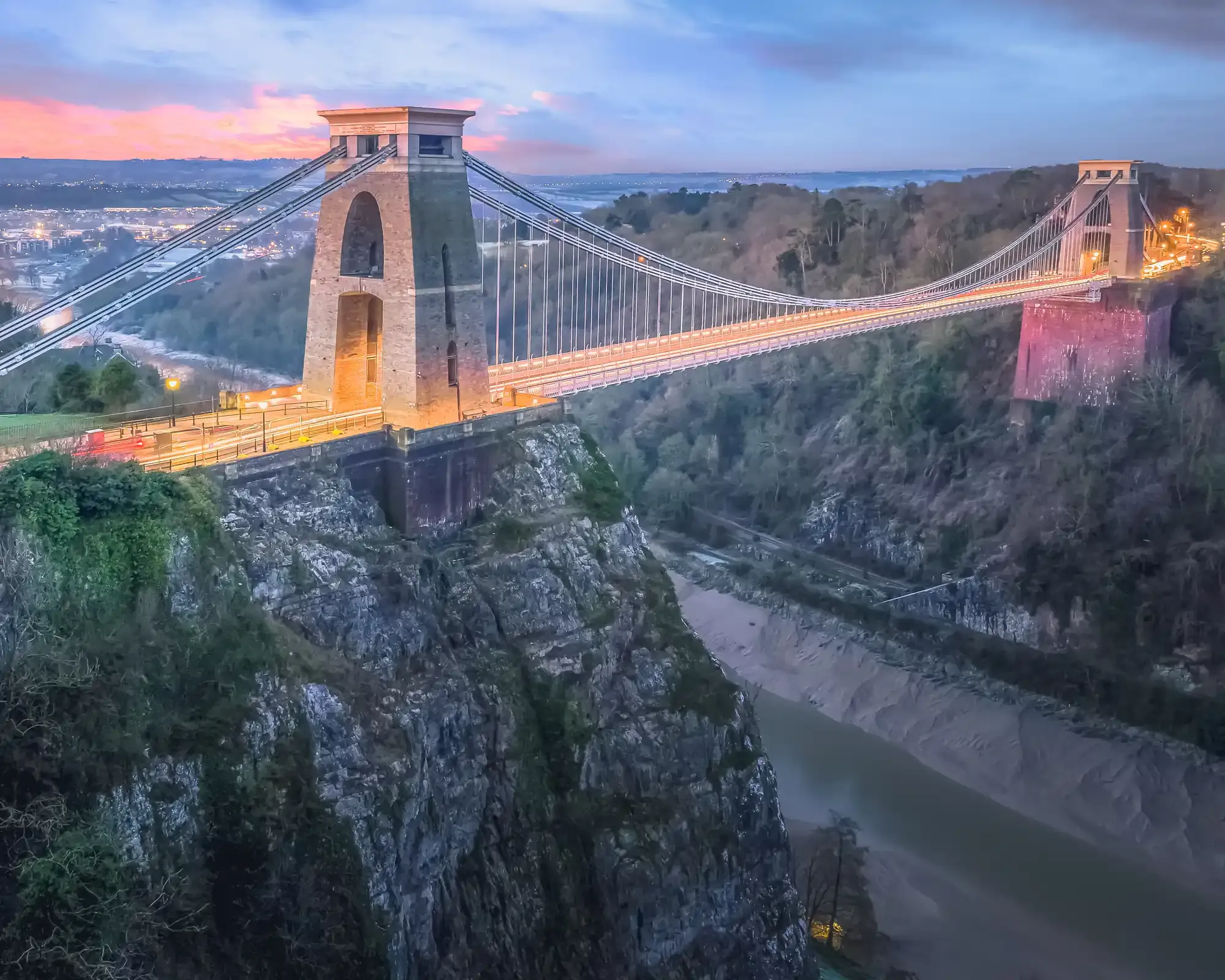 Clifton Suspension Bridge in Bristol at dusk, lit up across the Avon Gorge under a pink and blue sky.