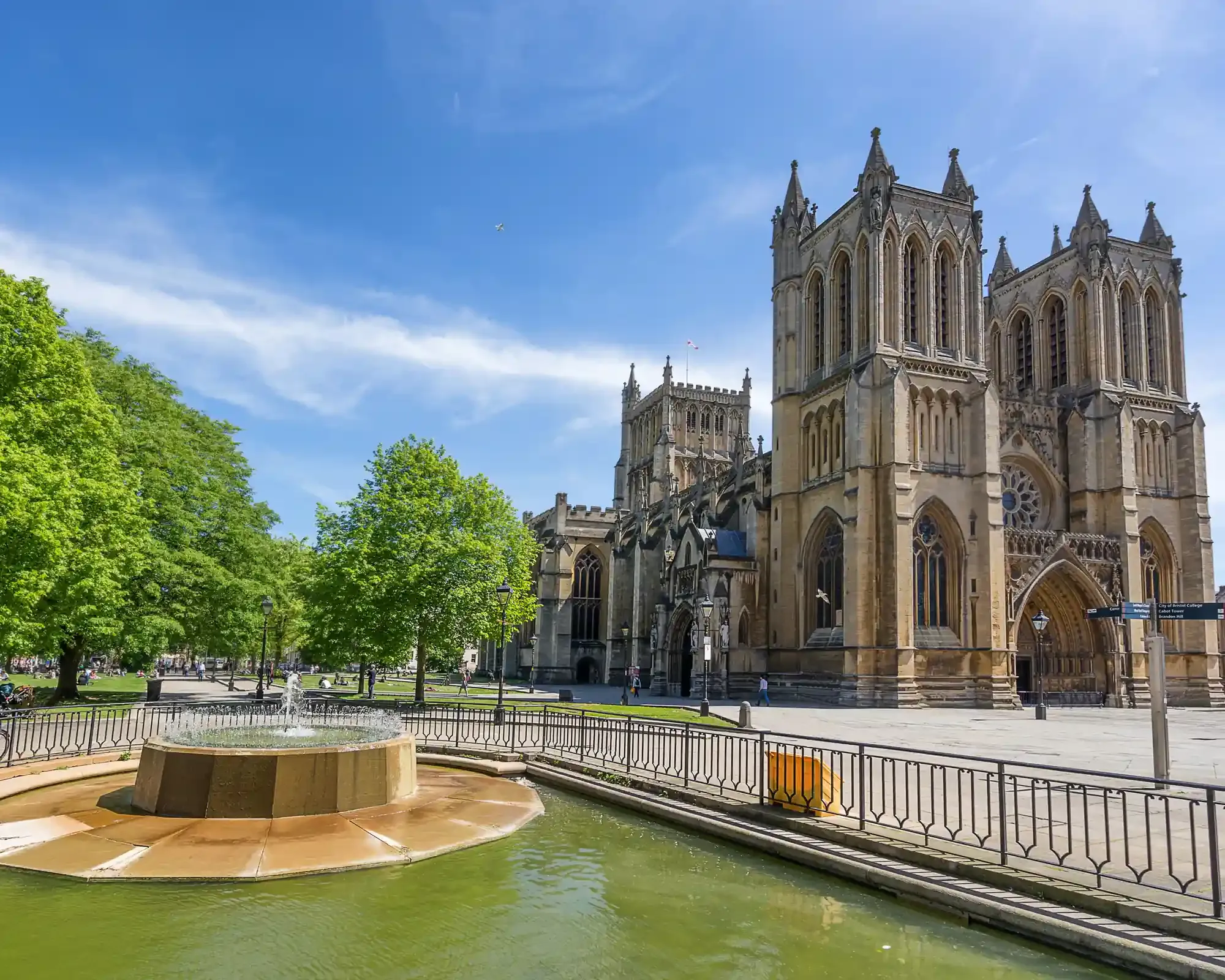 Bristol Cathedral on a sunny day, seen from College Green, with a fountain and leafy trees in the foreground.