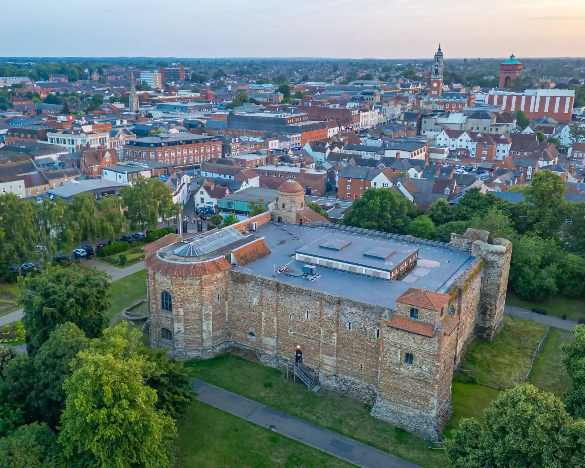 Aerial view of Colchester Castle’s Norman keep in Castle Park, with the Town Hall clock tower and Jumbo water tower on the city skyline.