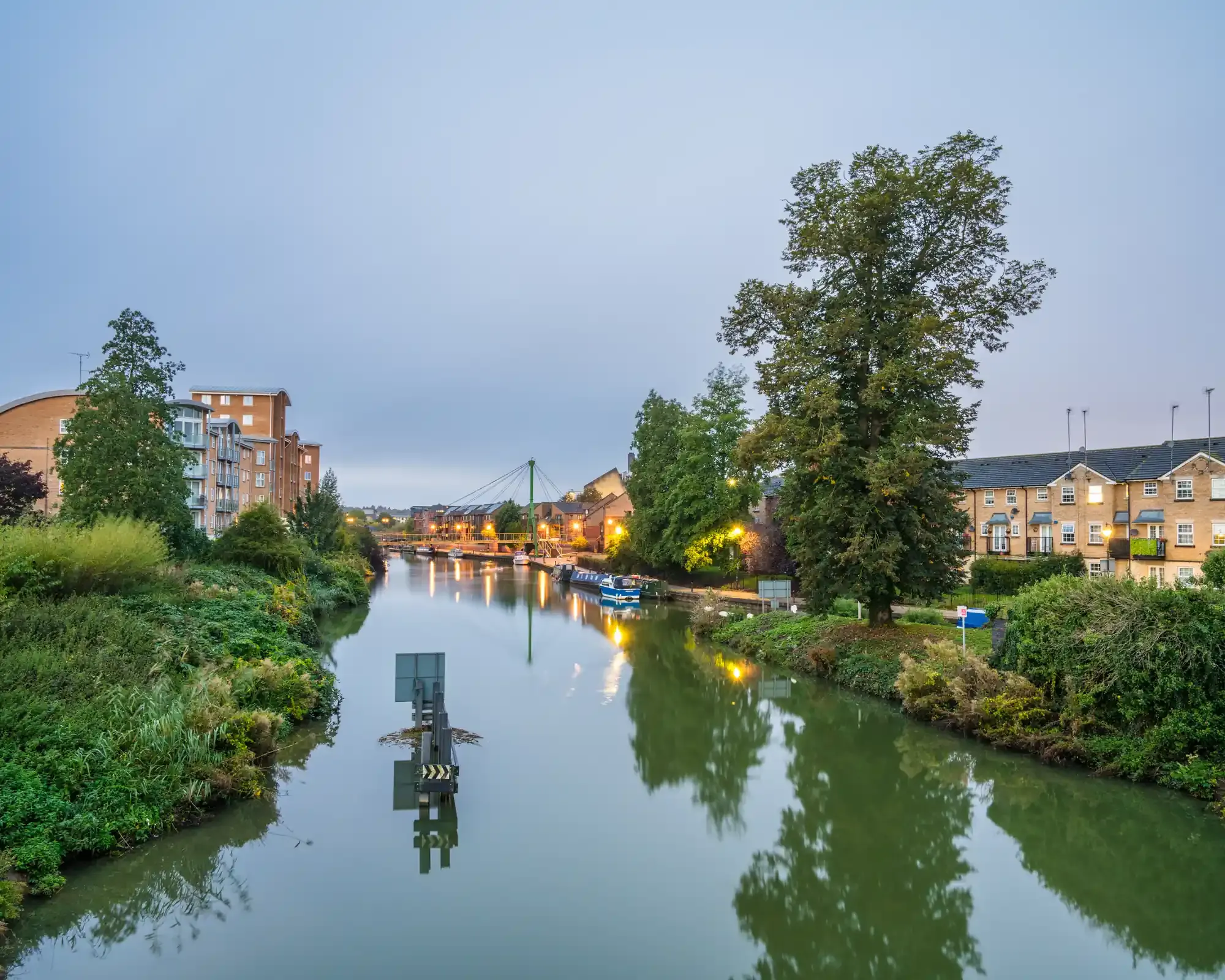 Riverside apartments and townhouses along the River Nene in Northampton at dusk, with narrowboats moored by a footbridge.