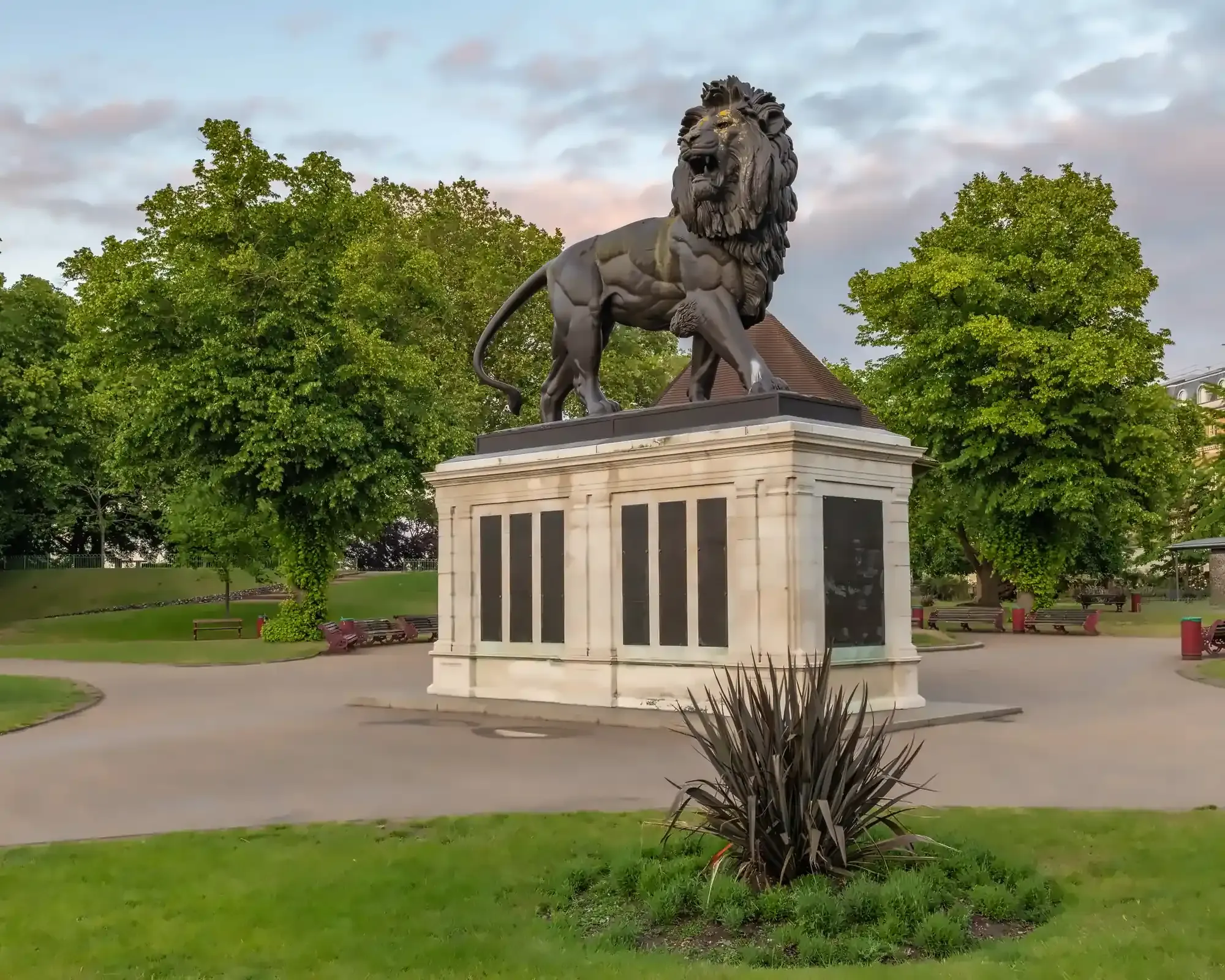 The Maiwand Lion statue in Forbury Gardens, Reading, standing on a stone memorial plinth surrounded by leafy trees and park paths under a soft evening sky.