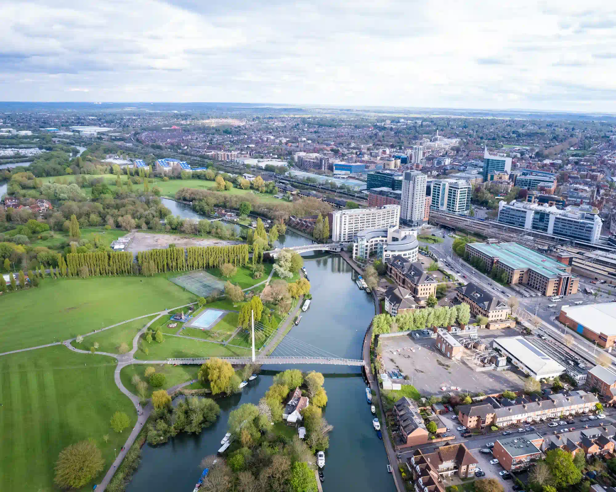 Aerial view of Reading, with the River Thames and Christchurch Bridge running between green parkland and the town centre, under a cloudy sky.