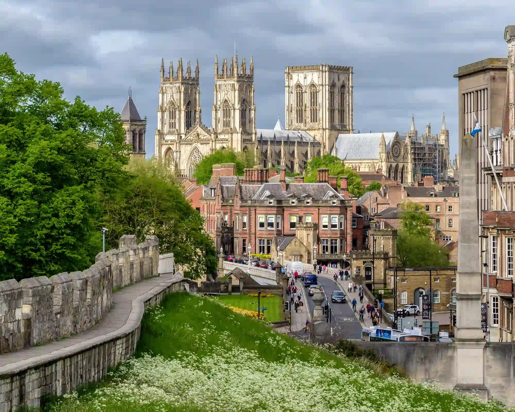 York Minster's towers rising above the city, seen from the medieval city walls, with a stone walkway, wild white flowers and a busy street below a cloudy sky.